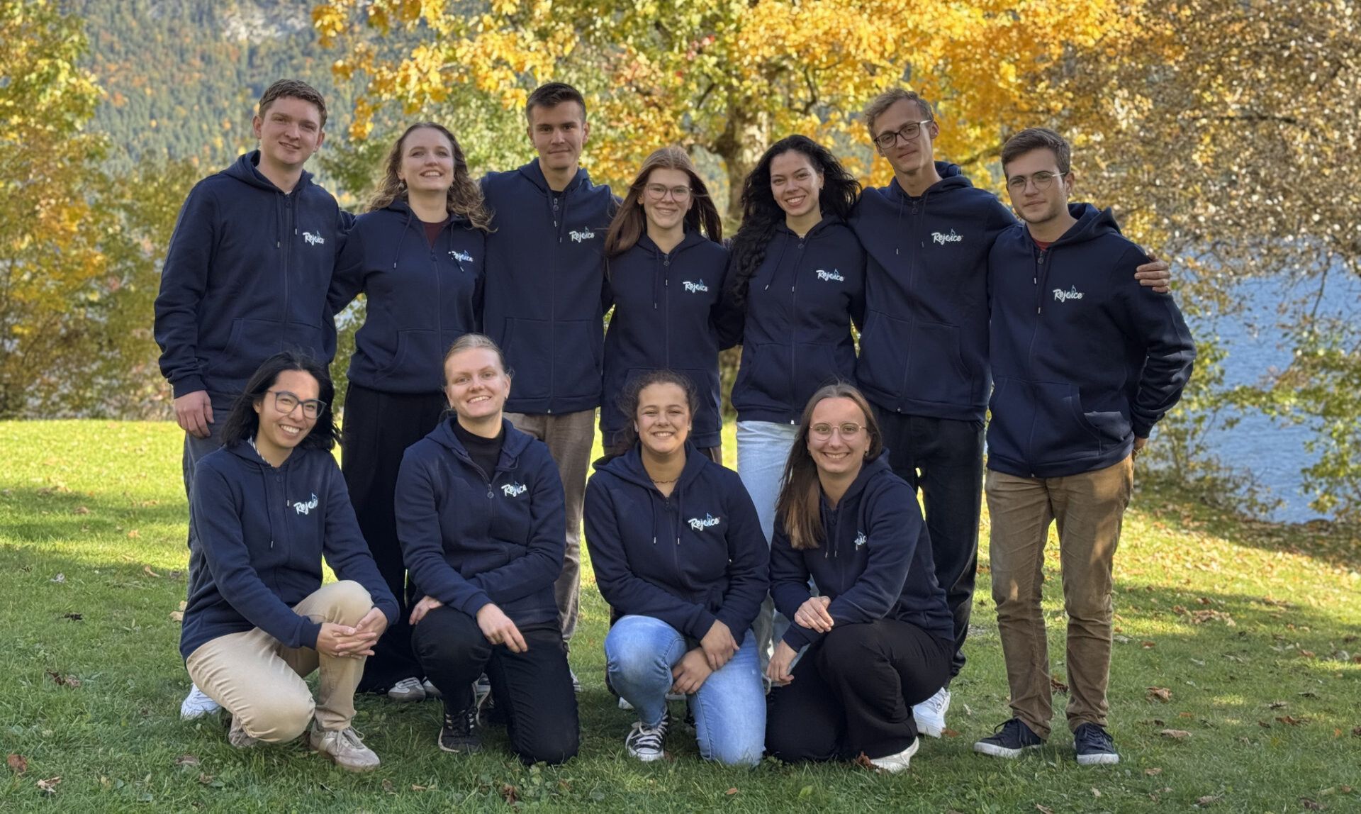 Die Rejoice-Gruppe 2025/26 in dunkelblauen Rejoice-Hoodies auf einer Wiese an einem See im Herbst