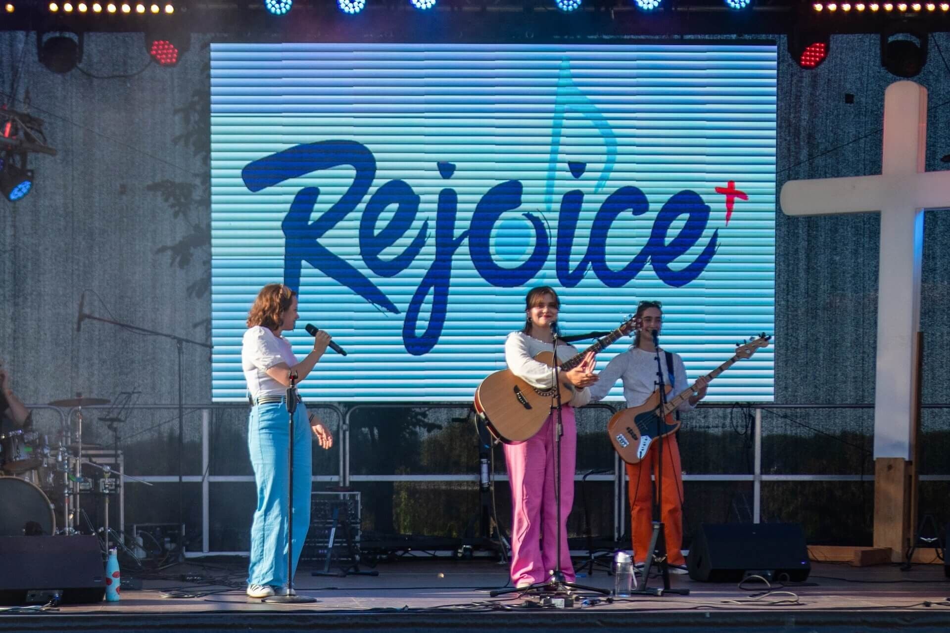 The Rejoice band on an open-air stage in front of a big screen showing the Rejoice logo